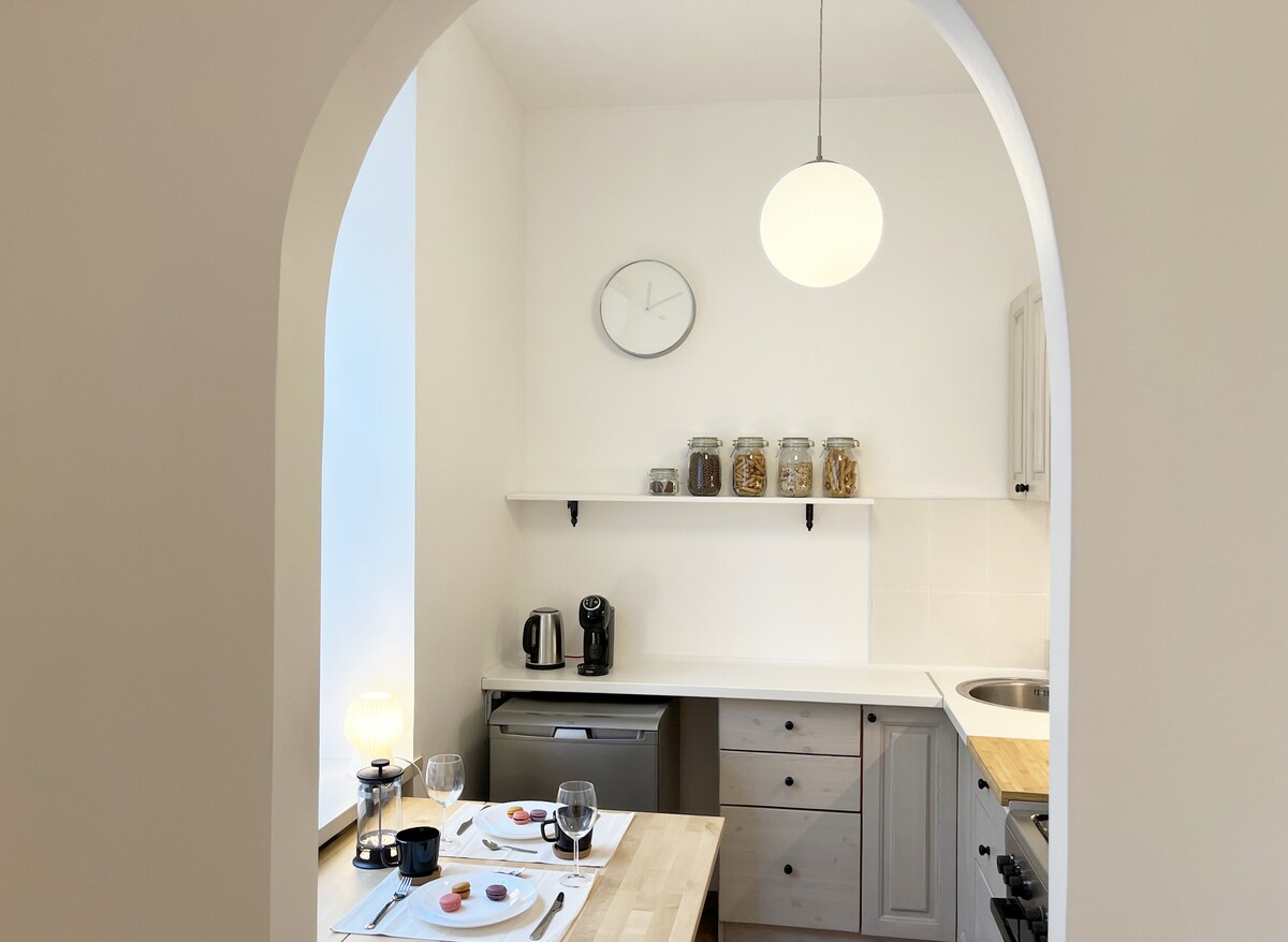 A light kitchen area is highlighted by a round pendant light and an archway. A wooden dining table is set for two with plates and glassware. A coffee machine and a selection of jars filled with snacks are visible on the countertop, complemented by modern appliances.