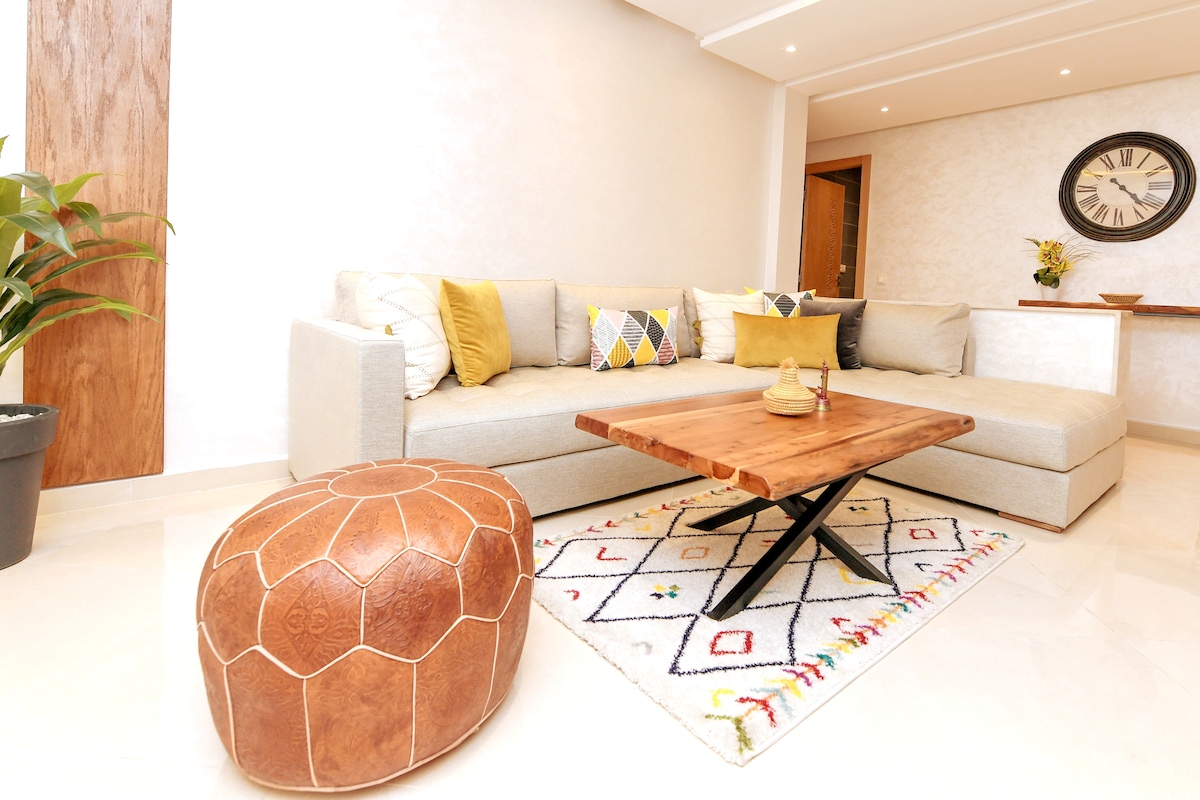 A spacious living area features a light-colored sectional sofa adorned with decorative cushions. A wooden coffee table sits atop a colorful patterned rug. A round leather pouf adds to the seating options, and a plant is positioned in one corner to enhance the ambiance.
