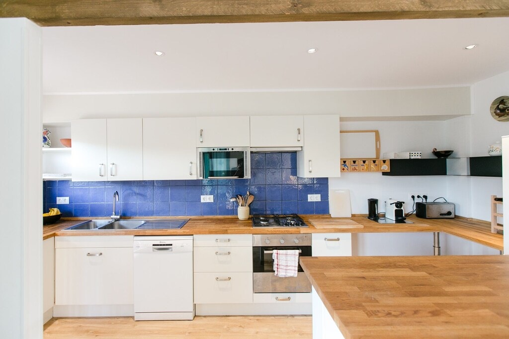 A modern kitchen is displayed, featuring white cabinetry and a blue tiled backsplash. Stainless steel appliances, including an oven and dishwasher, are visible. A wooden countertop provides ample workspace, accompanied by various kitchen utensils and appliances neatly arranged on shelves.