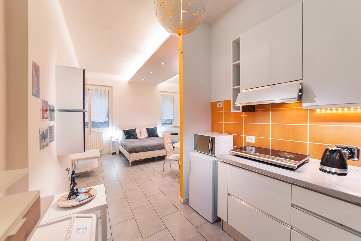 An open-plan living space features a well-equipped kitchen with modern appliances, including a microwave and kettle. The cozy sleeping area contains a spacious bed positioned against a softly lit orange accent wall. Natural light filters through sheer window curtains, enhancing the contemporary design.