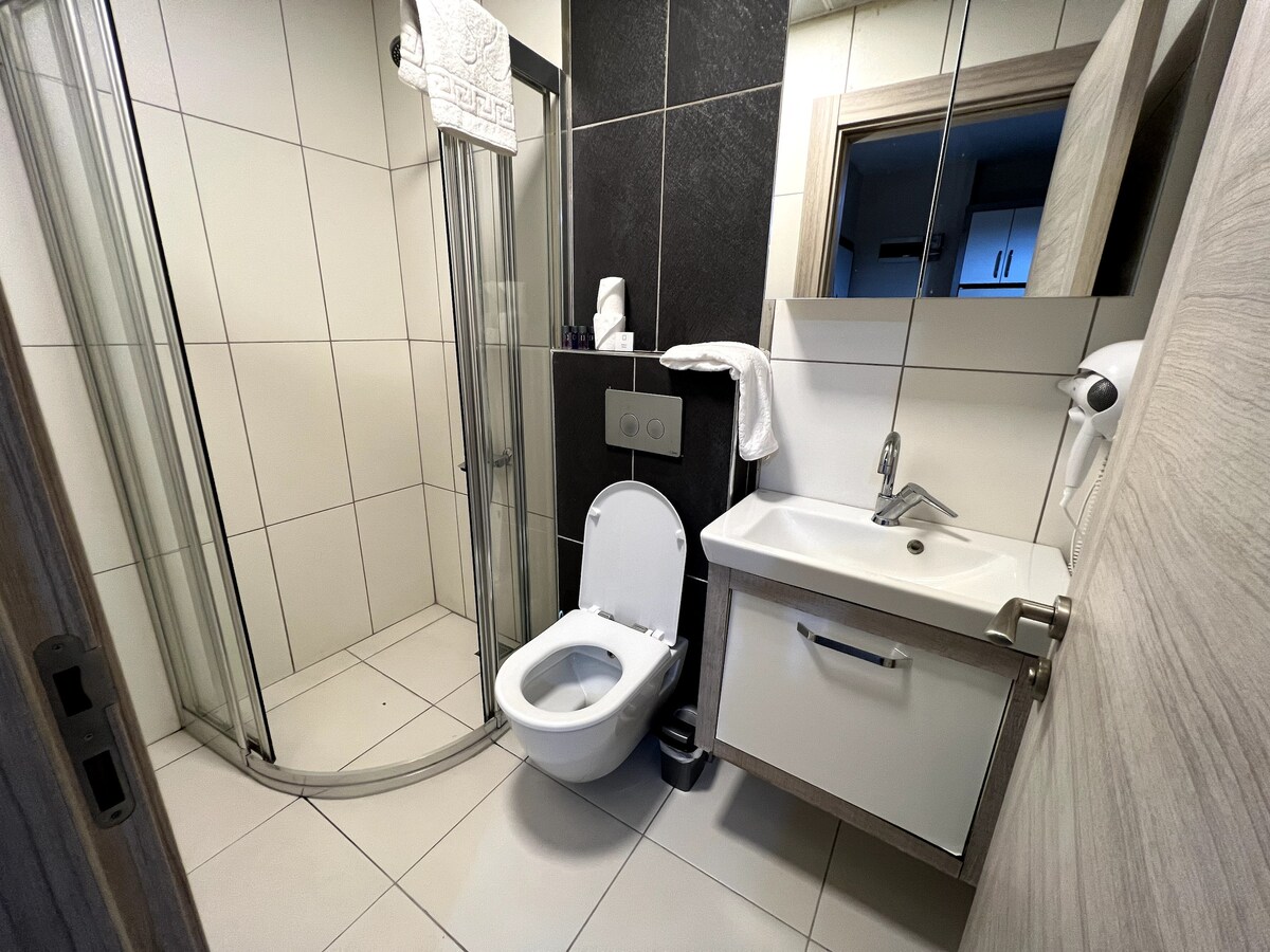 A modern bathroom space is presented, featuring a walk-in shower with glass doors, a toilet, and a sleek vanity with a sink. Neutral tiles line the walls and floor, creating a clean and functional atmosphere. Complimentary toiletries are neatly arranged for convenience.