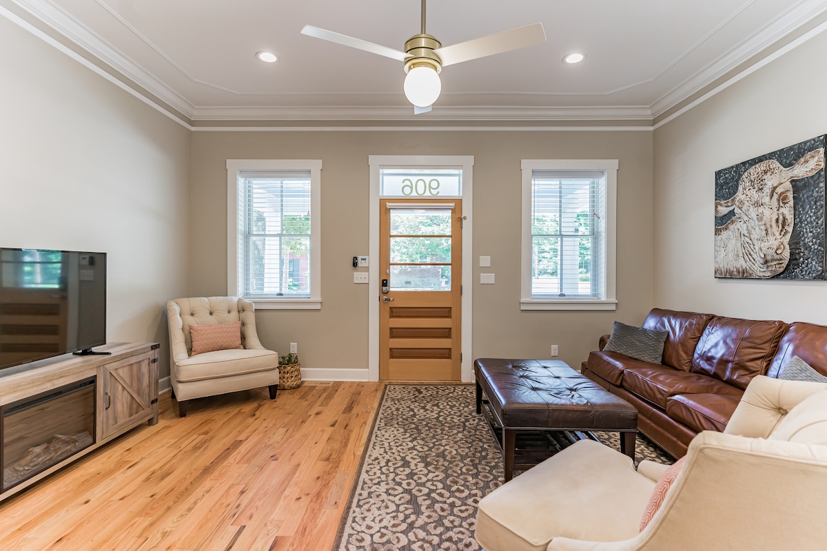 A welcoming living area features a combination of light hardwood floors and neutral walls. A comfortable leather sofa faces a wooden media console below a flat-screen television. Natural light enters through two windows, while a ceiling fan adds to the fresh ambiance of the space.