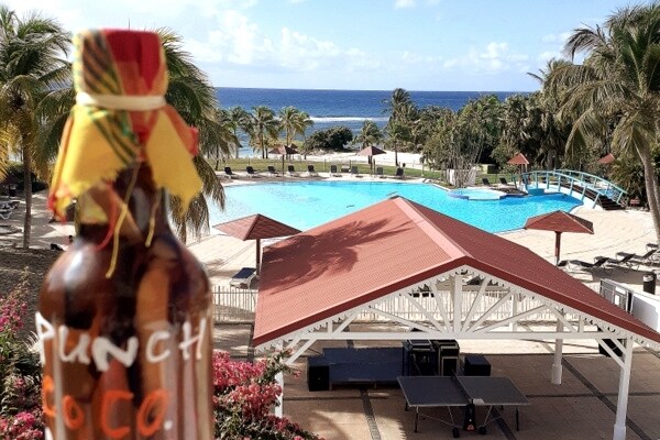 A clear view of a swimming pool is presented, surrounded by palm trees and lounge chairs. A beachfront extends in the background, with the ocean visible under a blue sky. A bottle of Punch Coco is positioned in the foreground, adding a local touch to the scene.