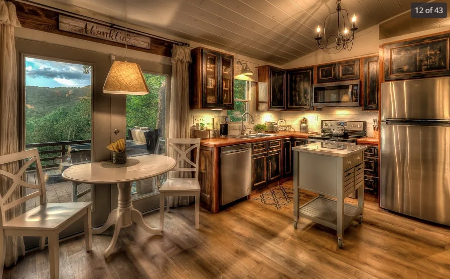 A well-equipped kitchen features dark wood cabinets, stainless steel appliances, and a small island. A round dining table with two chairs is positioned by large glass doors, which offer views of the surrounding greenery. A pendant light illuminates the space, creating a cozy atmosphere.