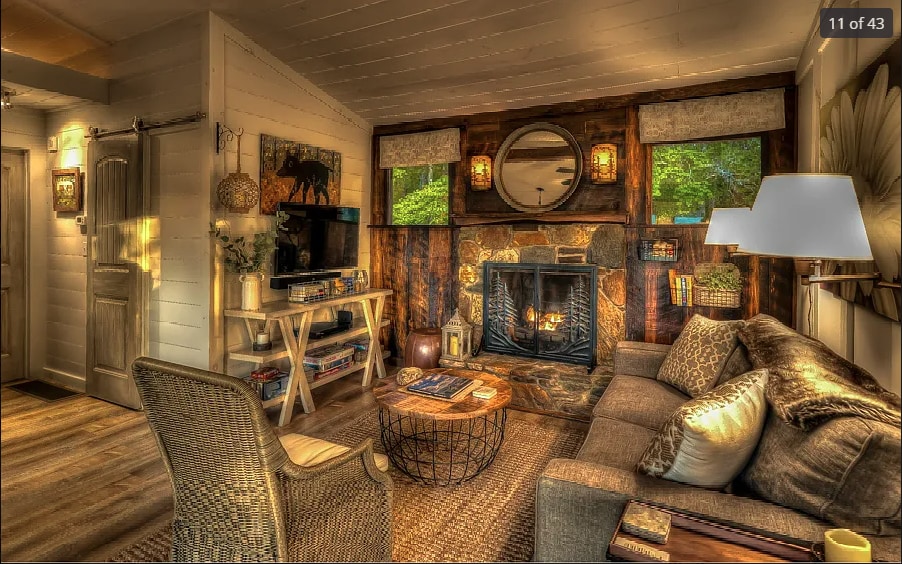 A welcoming living area is presented, featuring a comfortable sofa with textured cushions and a coffee table in front. A stone fireplace is centered against the wood-paneled wall, while large windows bring in natural light, offering views of the outdoors. Decorative elements enhance the cozy atmosphere.
