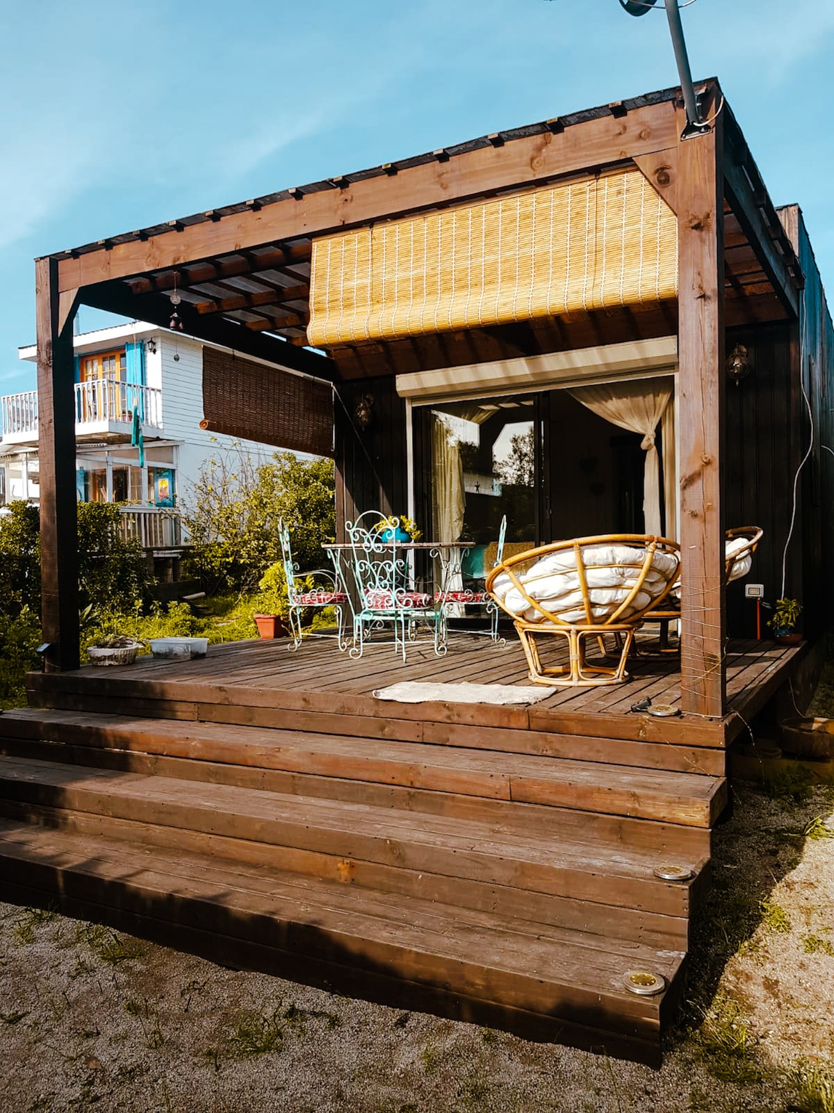 An outdoor deck is shown with wooden steps leading up to a covered area. A table and colorful chairs are arranged under a bamboo shade. Lush greenery surrounds the space, creating a private and inviting environment.