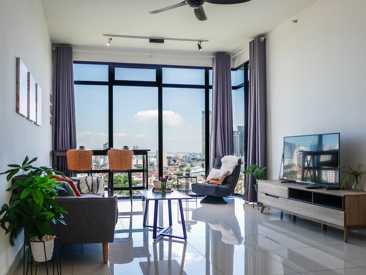A modern living area features large windows offering city views. Comfortable seating is arranged around a coffee table, while a television and decorative plants enhance the ambiance. Neutral tones and natural light create a spacious and inviting environment.