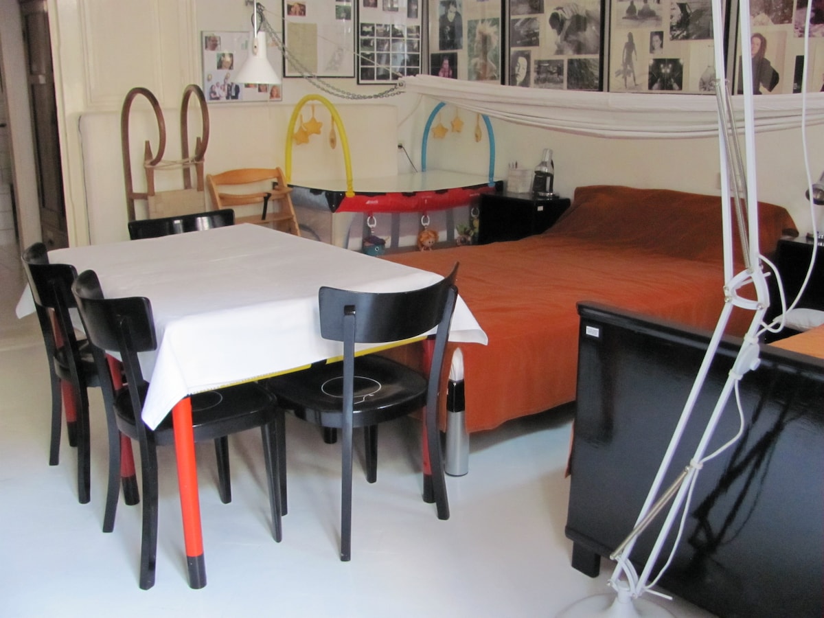 A cozy living area features a spacious bed dressed in an orange coverlet. A dining table surrounded by six black chairs is positioned nearby. The walls display an array of photographs, while various furniture pieces add a touch of character to the space.