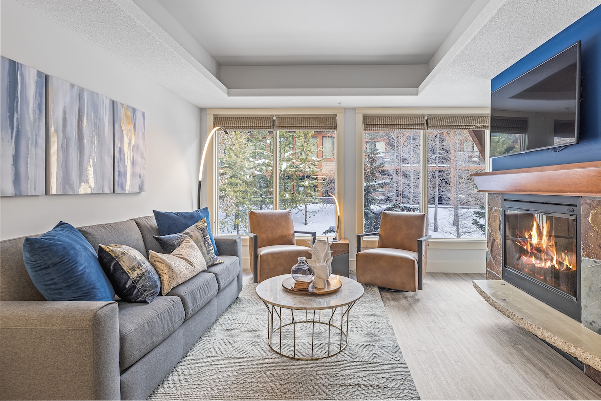 A cozy living area is highlighted by a modern fireplace, with a spacious gray sofa and two brown armchairs arranged around a circular coffee table. Large windows allow natural light and offer views of the outdoor scenery, enhancing the room's inviting ambiance.