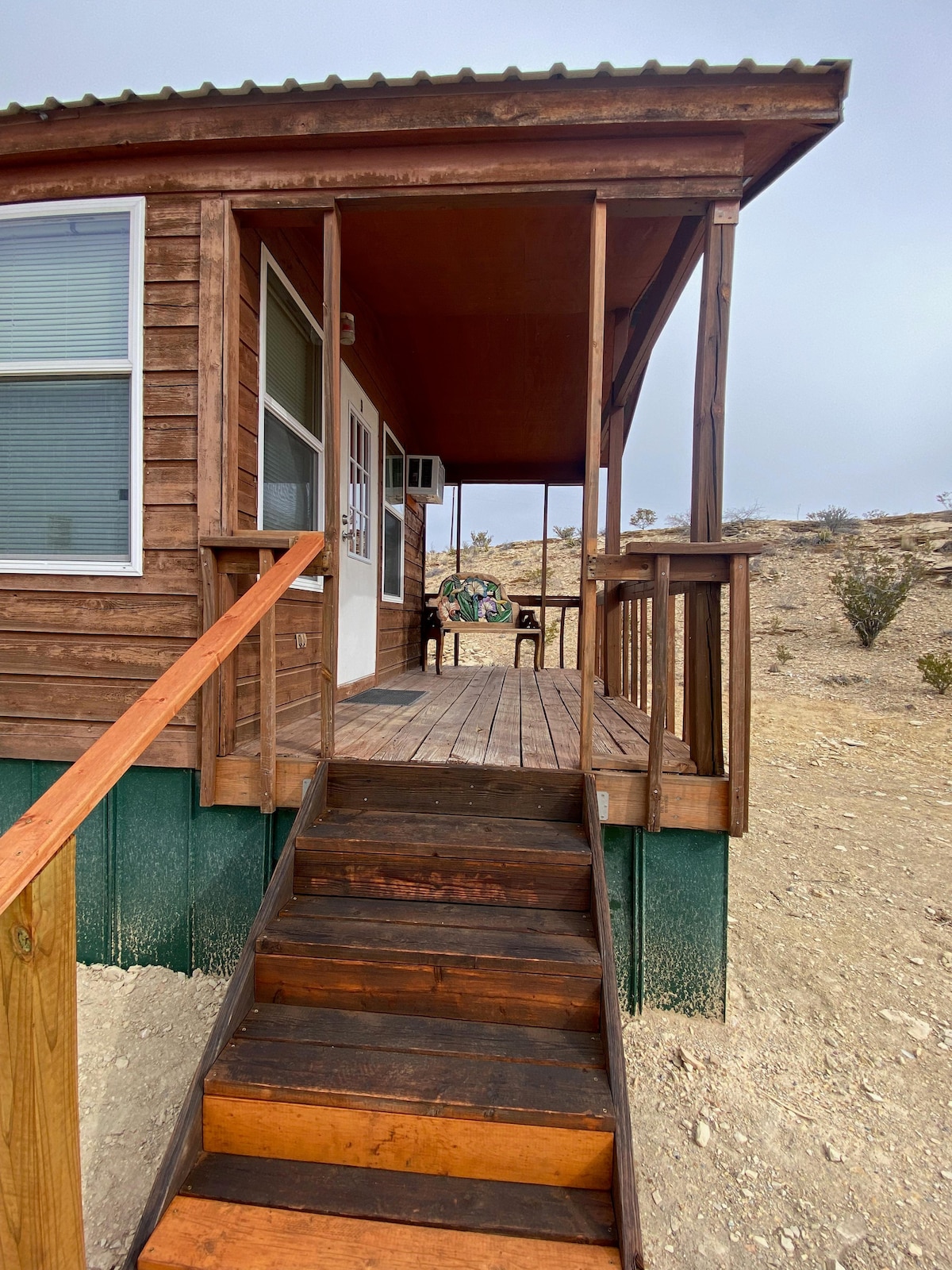 A wooden porch with steps leads to a front entrance of the Loft, featuring large windows and a seating area. The porch is surrounded by a natural landscape, providing a connection to the outdoor environment.