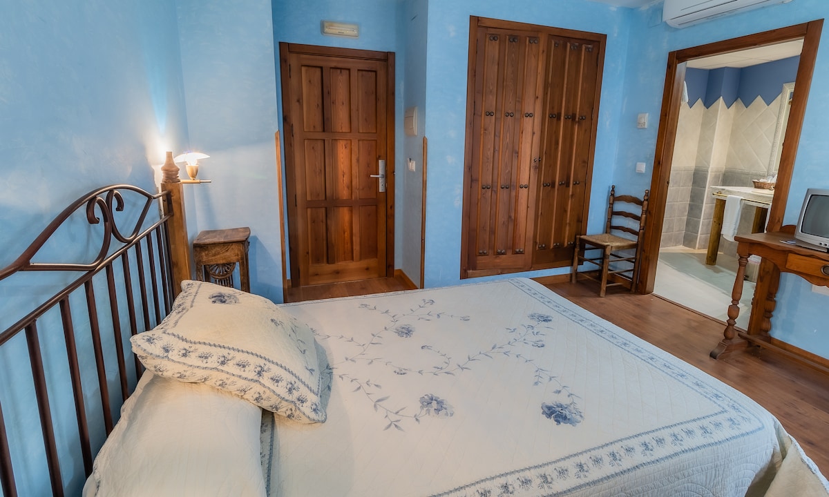 A light blue room features a metal-frame bed with decorative bedding and a simple floral design. A wooden door and wardrobe are visible, along with a chair. A small wooden table stands beside the bed, and a partially open door leads to a bathroom.