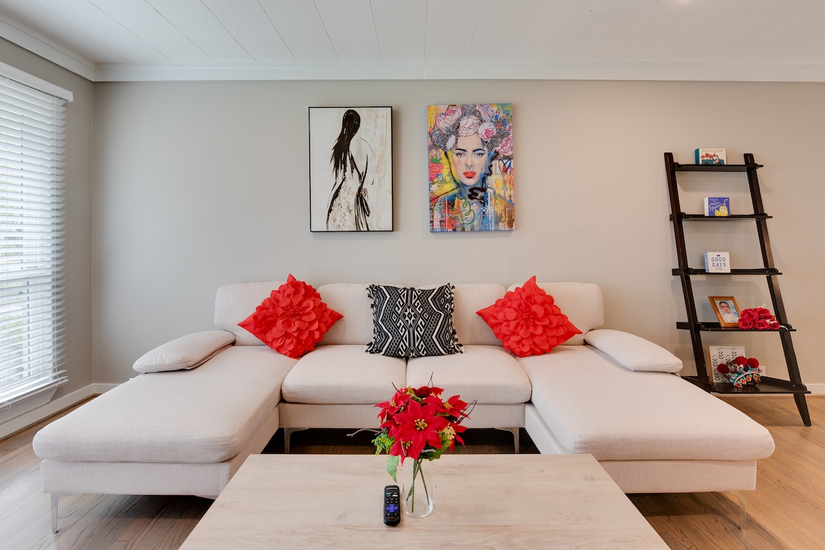A modern living area features a light-colored sectional sofa adorned with red decorative pillows. A coffee table with a floral arrangement sits in front. Colorful artwork is displayed on the walls, and a stylish shelving unit holds various decorative items.