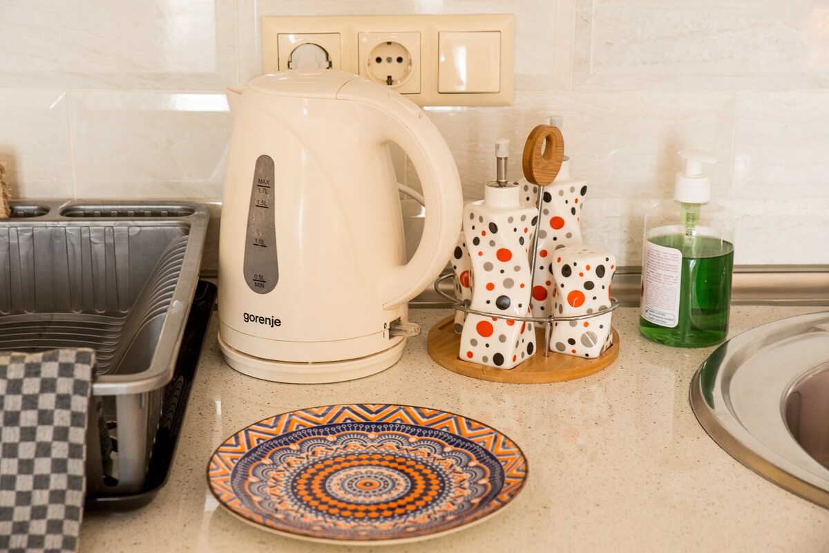 A sleek electric kettle is positioned beside a neatly arranged drying rack and a decorative plate featuring intricate patterns. A set of patterned mugs is displayed on a wooden holder, with a liquid soap dispenser and sink visible in the background.