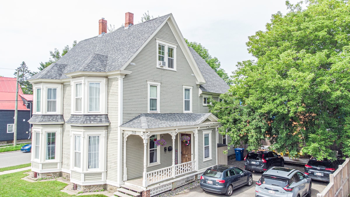 The image depicts the historic Harvey House from an elevated angle, highlighting its charming architecture and well-maintained exterior. The two-story building features bay windows and a wraparound porch. A driveway is visible, accommodating several vehicles, with lush trees providing shade nearby.
