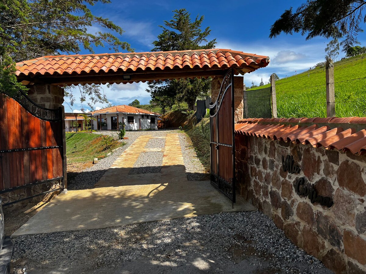 A welcoming entrance is revealed through a rustic stone archway topped with a terracotta roof. A driveway lined with gravel leads towards a lush green landscape, framed by trees. The house is visible in the distance, blending harmoniously with the surrounding nature.