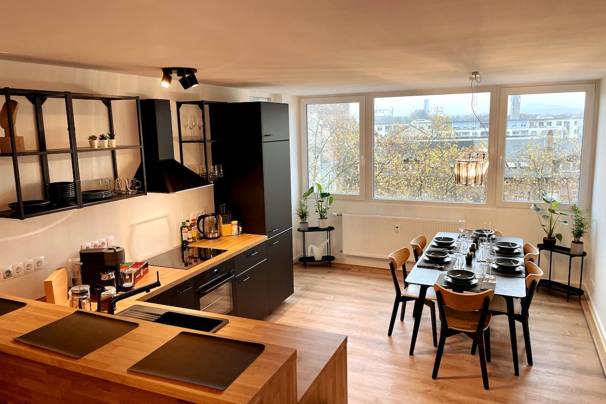 A modern kitchen and dining area is highlighted by large windows, providing ample natural light. The well-equipped kitchen features sleek black cabinetry and a wooden countertop. A dining table is set for six, surrounded by wooden chairs, with greenery enhancing the inviting atmosphere.