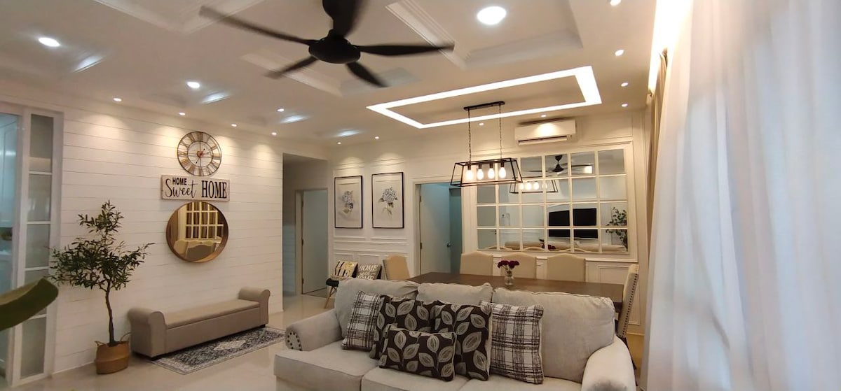 A bright living area is showcased, featuring a spacious sofa adorned with patterned cushions. A ceiling fan circulates air, while a large clock and framed artwork add character. Natural light filters through sheer curtains, illuminating the elegant dining area in the background.
