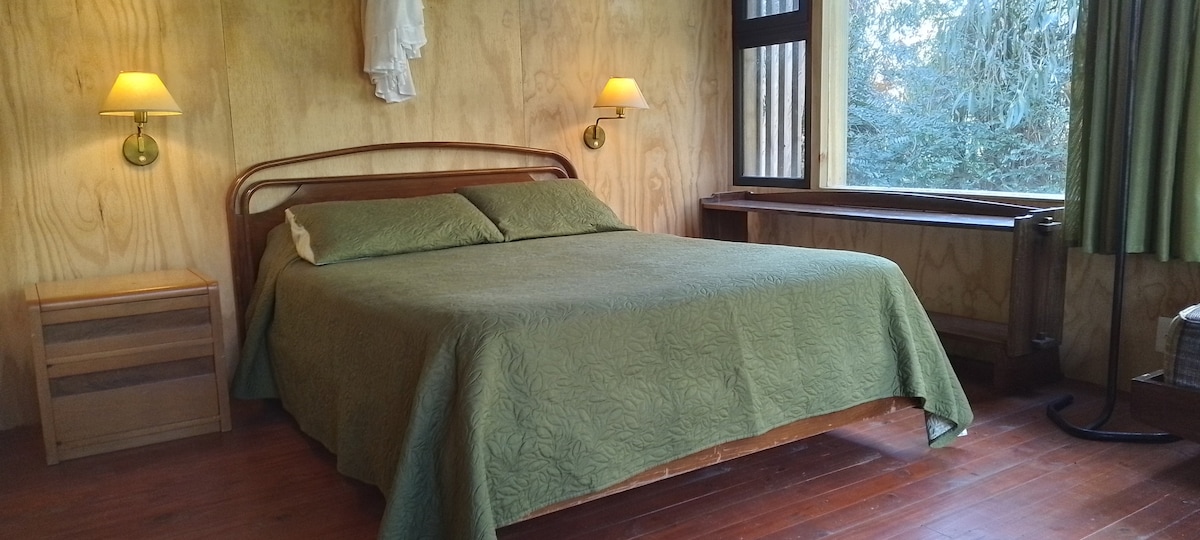 A modern bedroom features a spacious bed with a green coverlet. Two bedside lamps provide soft lighting, and a large window offers views of the surrounding trees. Wooden walls and floors create a natural ambiance, enhancing the room's minimalist design.