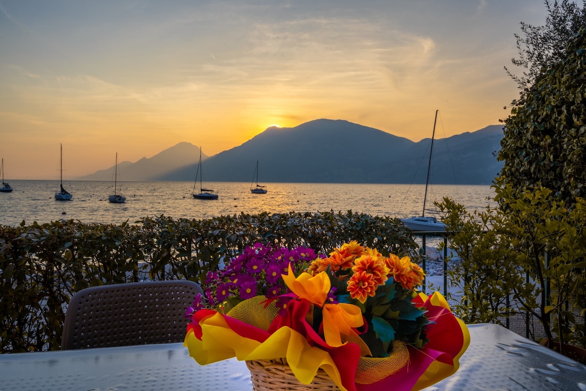 A table is set outdoors featuring a vibrant floral arrangement in a basket, positioned in front of the tranquil waters of Lake Garda. The scene is illuminated by a warm sunset, with silhouettes of mountains and sailboats visible in the background.