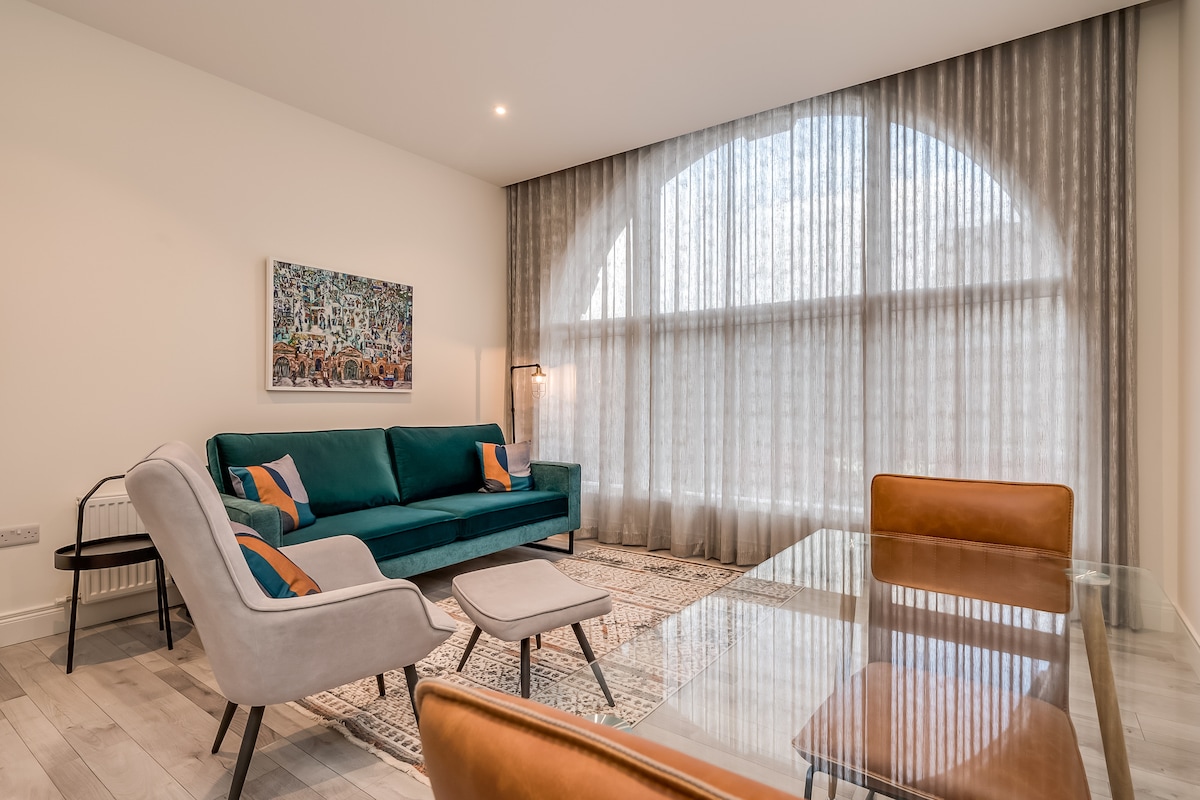 A modern living area features a teal sofa, two light gray armchairs, and a glass dining table. An artwork hangs on the wall, complementing the light streaming through a large floor-to-ceiling arched window covered by sheer curtains. A neutral-colored rug adds warmth to the space.