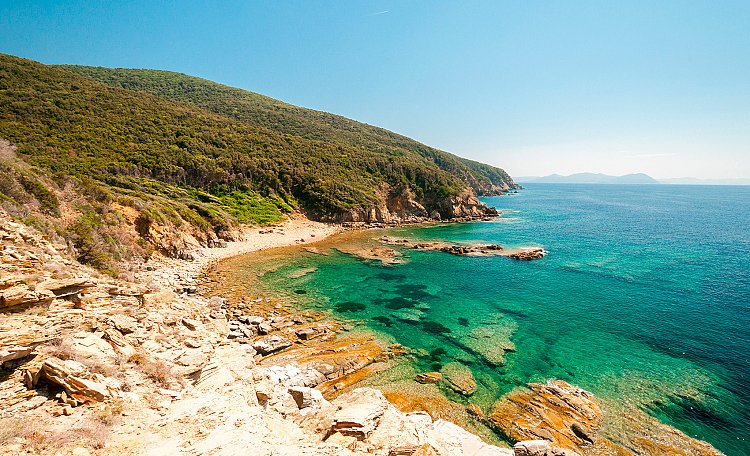 The image captures a tranquil coastal landscape, featuring clear turquoise waters gently lapping against rocky shores. Lush green hills frame the scene, leading toward the horizon where distant islands are visible under a bright blue sky.