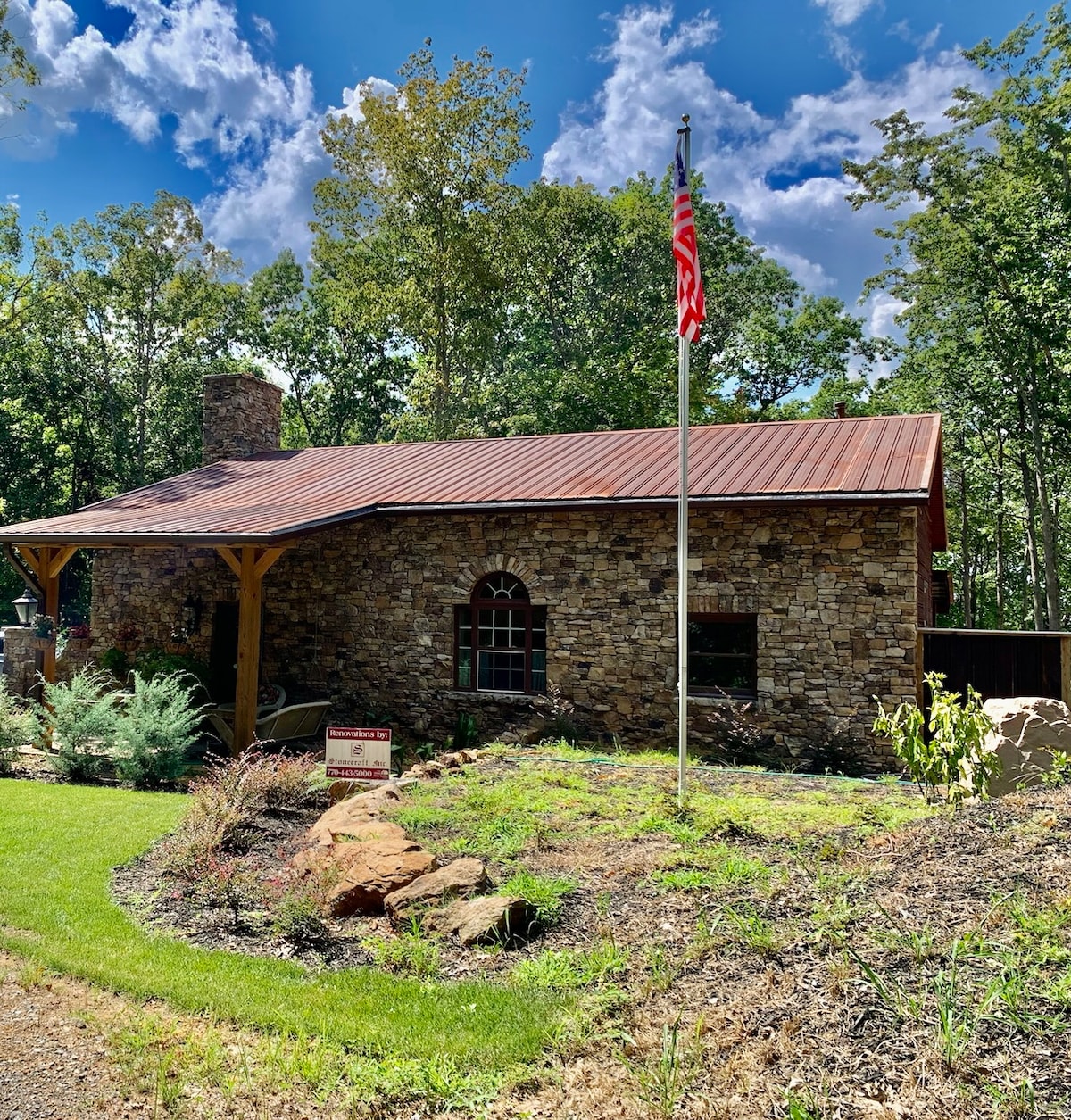 A rustic stone house is showcased against a backdrop of lush trees and a bright blue sky. An American flag is planted at the front, and a wooden porch with a sloping roof frames the entrance, complementing the natural surroundings.