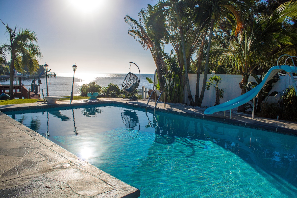 A serene pool area is framed by lush palm trees and overlooks a tranquil bay. The inviting water reflects sunlight, while a spiral slide and a hanging chair add unique touches for leisure. The space is designed for relaxation and enjoyment of the scenic ocean view.