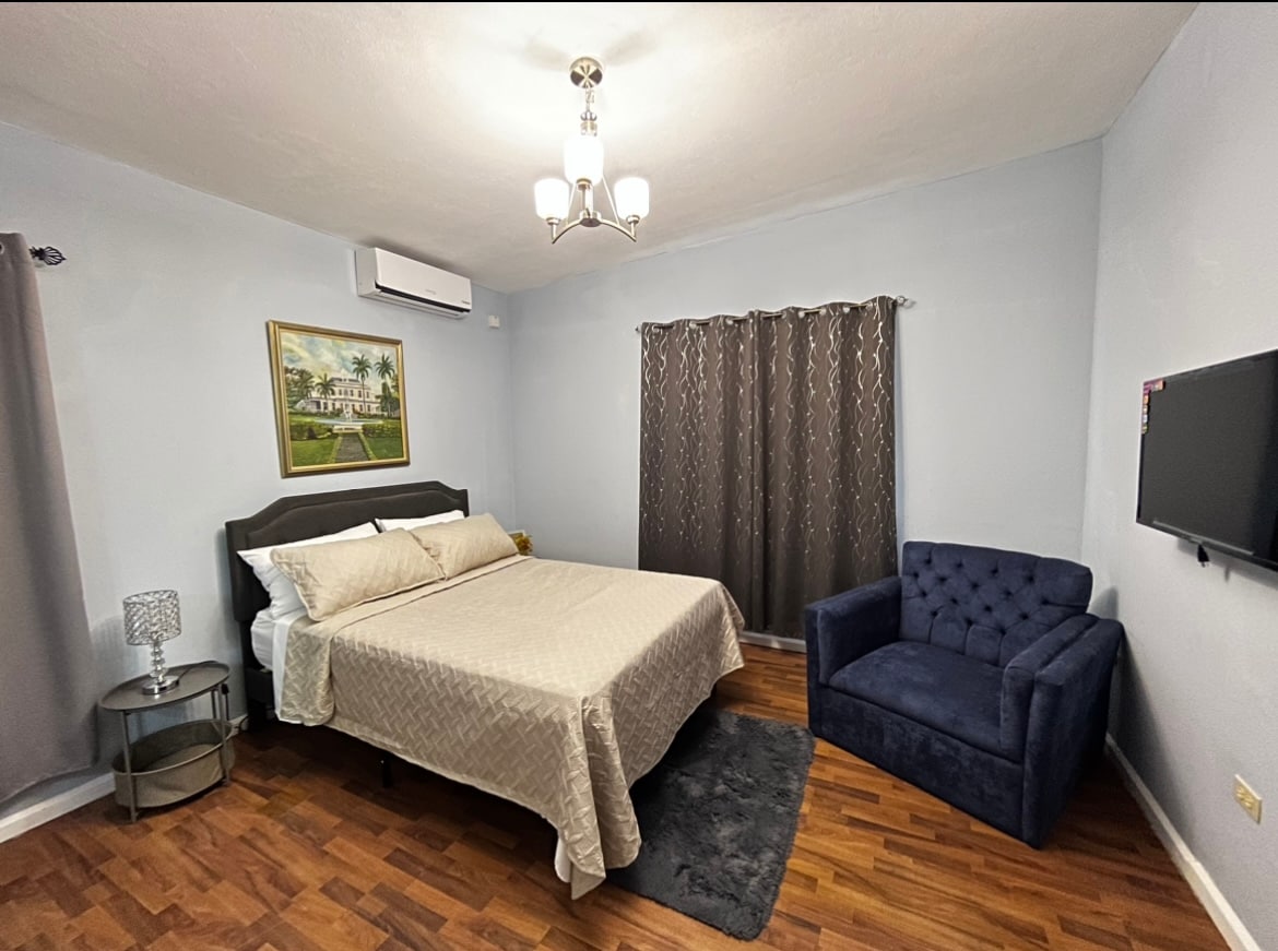 A cozy room features a spacious bed dressed in neutral linens. A blue upholstered chair offers additional seating, while a round side table sits nearby. Light wood flooring complements the soft blue walls, and natural light filters through a window covered with patterned curtains.