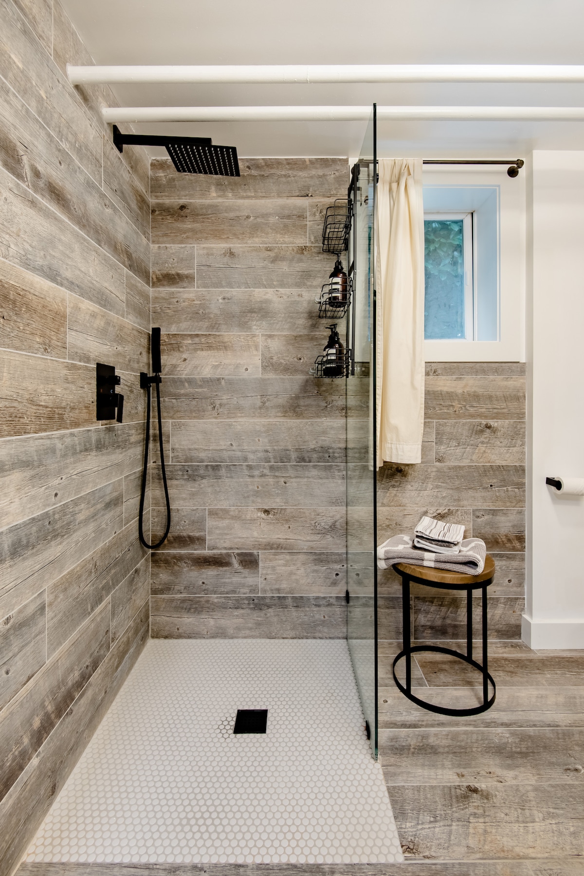 A modern shower area features a glass panel and a rainfall showerhead. Textured wooden walls are complemented by a patterned tile floor. A compact stool is positioned nearby, adorned with neatly arranged towels, while natural light filters through a small window.