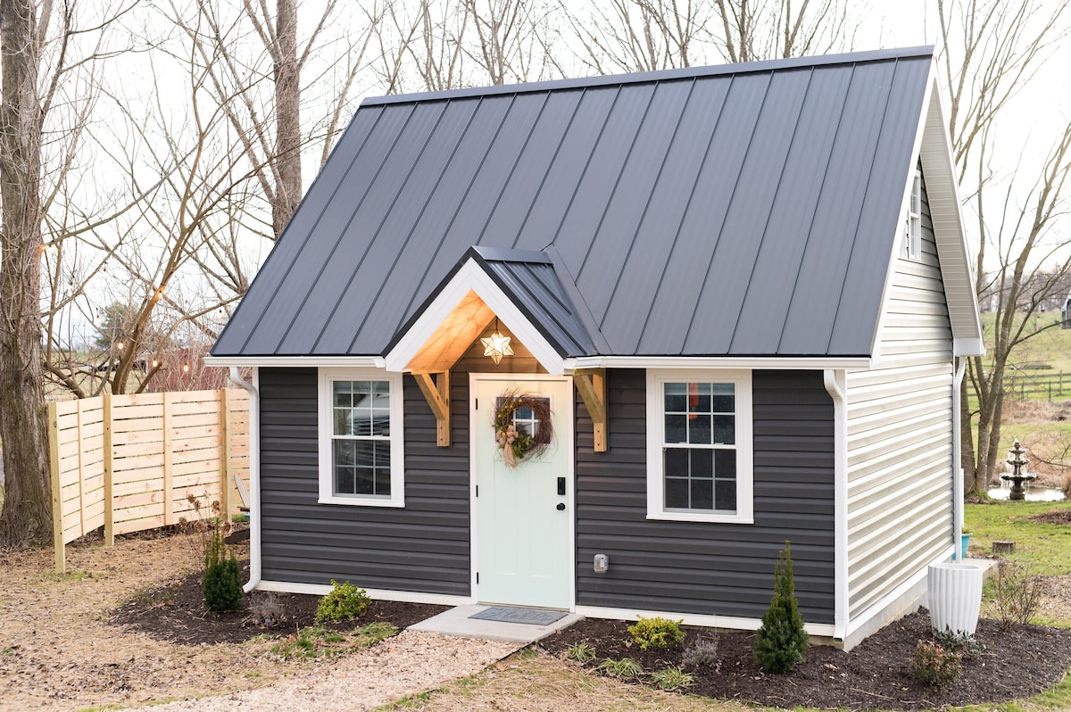 The exterior of the springhouse is shown, featuring a modern design with a sloped metal roof and horizontal siding. The entrance is framed by wooden features and adorned with a seasonal wreath. Landscaping includes small shrubs and a gravel pathway leading to the door.