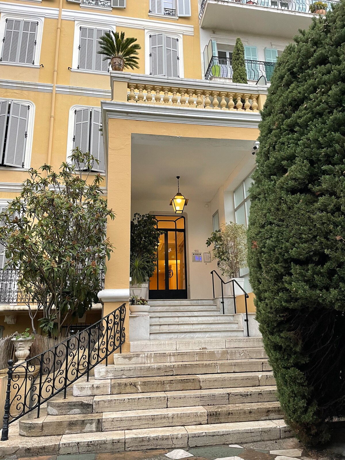 An entrance to a beautifully maintained building is highlighted by a series of steps leading to an elegant door. Flanked by greenery and adorned with a lantern, the inviting facade features yellow walls and decorative shutters. A mix of natural and architectural elements is visible.
