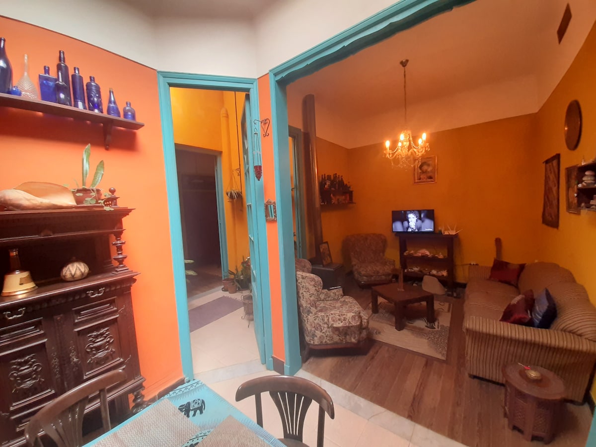 An open entryway leads into a vibrant lounge area with warm yellow walls. Comfortable seating arrangements, including floral-patterned armchairs and a striped sofa, surround a wooden coffee table. A vintage cabinet stands against the wall, while a chandelier adds soft illumination to the space.