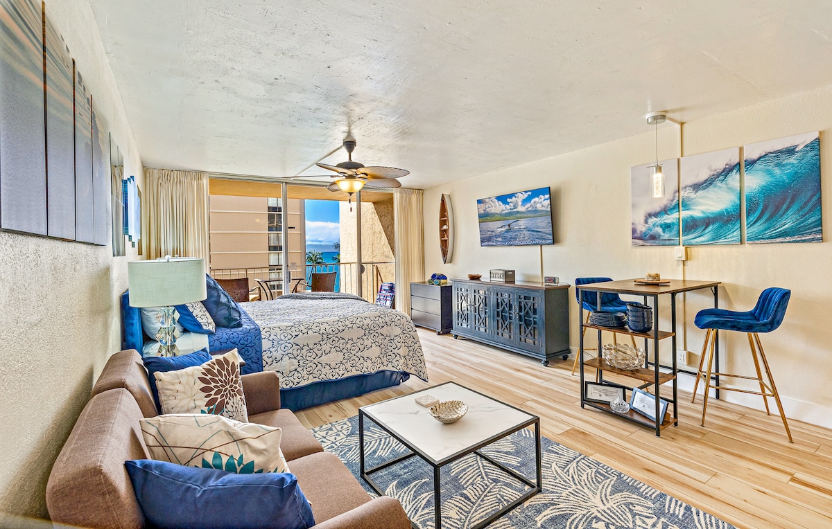 A spacious studio is showcased with wood flooring and natural light flowing in through large sliding glass doors leading to a lanai. The room features a king size bed, a seating area with a sofa, and a small dining table. Ocean views are visible.