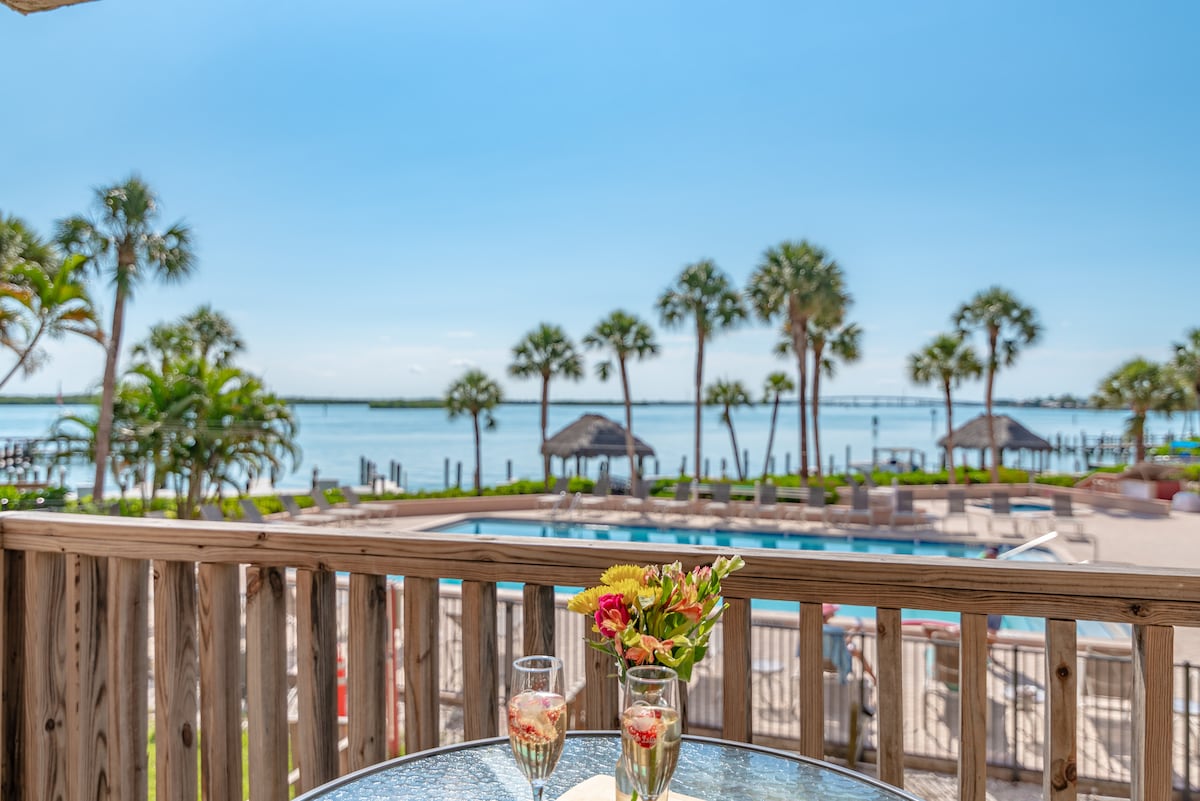 A serene view from the balcony features a table set with two glasses and a small floral arrangement. The background reveals palm trees, a swimming pool, and glistening water under a clear blue sky.