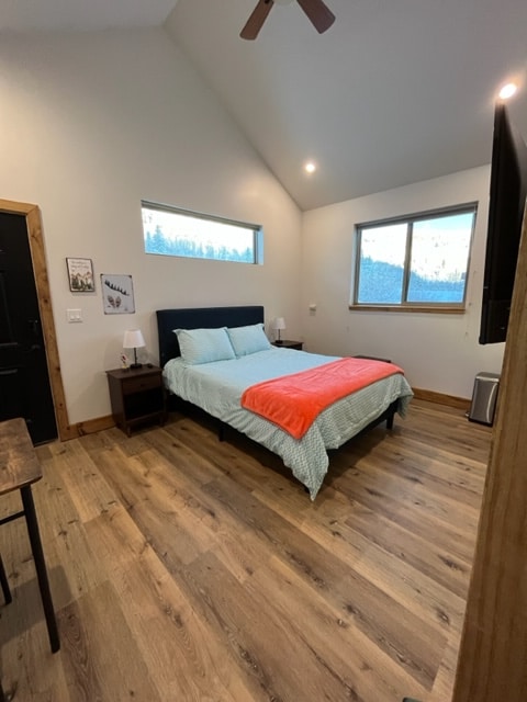 The room features a queen bed with a light blue quilt and a bright orange throw. There are two bedside tables with lamps, and a small table is positioned nearby. Large windows provide views of the surroundings, and warm wood flooring adds to the ambiance.