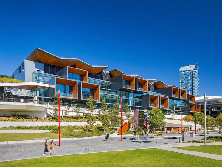 A modern building complex features a series of angular rooflines and large glass windows. The striking design is complemented by landscaped greenery and wide walkways. Tall trees and vibrant red poles provide visual interest against the backdrop of a clear blue sky.