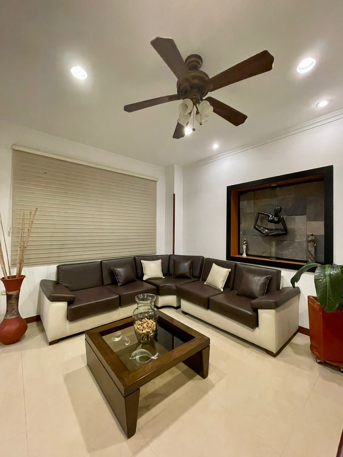 A comfortable seating area is furnished with a contemporary sectional sofa in brown and white. A glass coffee table sits in the center, complemented by decorative items. Soft lighting is provided by ceiling fixtures, highlighting the elegant decor and an artwork displayed on the wall.