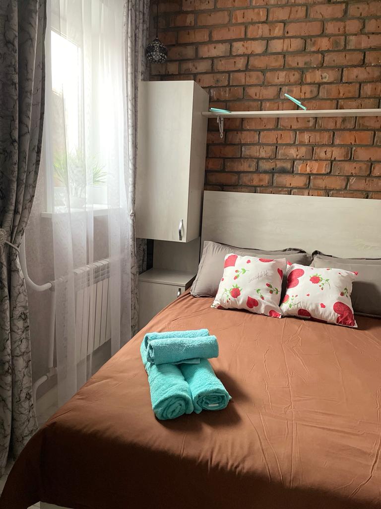 A cozy bedroom features a spacious bed adorned with brown bedding and decorative pillows. Freshly laundered towels are neatly arranged at the foot of the bed. Natural light filters through sheer curtains, illuminating the exposed brick wall and providing a warm ambiance.