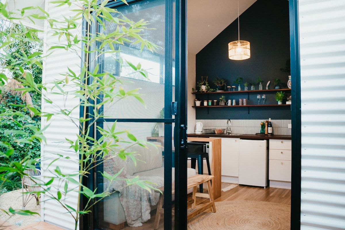 A welcoming entrance reveals a cozy living space. Natural light illuminates a kitchenette equipped with shelves, a sink, and appliances. A lounge area with a textured blanket invites relaxation, while plants frame the door, enhancing the connection to the vibrant outdoor setting.