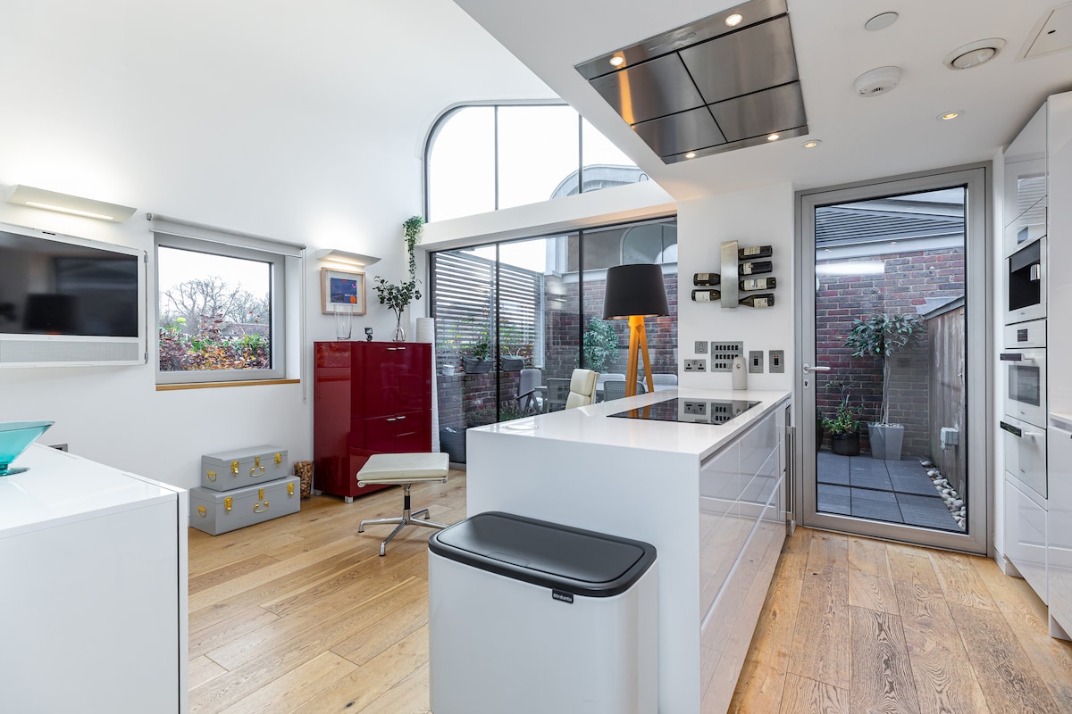 A modern kitchen features sleek white cabinetry complemented by a minimalist island. Natural light floods the space through large windows, creating an open feel. A stylish lamp sits atop the island, while a private terrace is visible through glass doors.