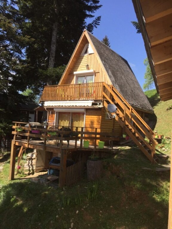 A charming chalet features a distinct A-frame design set against a backdrop of towering trees. A wooden deck wraps around the front, with steps leading to the entrance. Large windows allow natural light to fill the interior, while colorful flower pots add a touch of vibrancy.