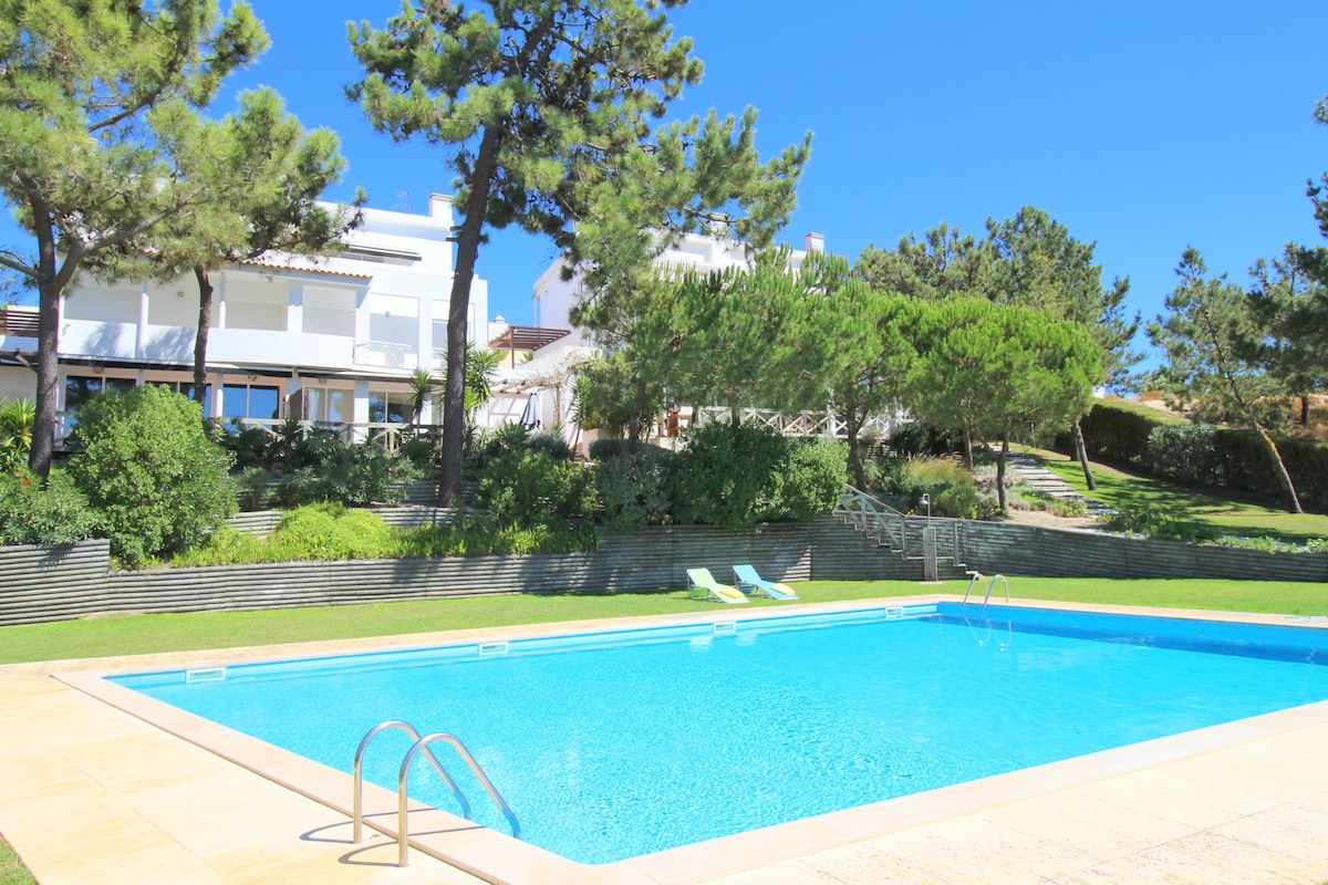 A clear blue swimming pool is surrounded by lush greenery and tall pine trees. Sun loungers are placed by the pool's edge. The villa can be seen in the background, with expansive windows reflecting the sunlight.