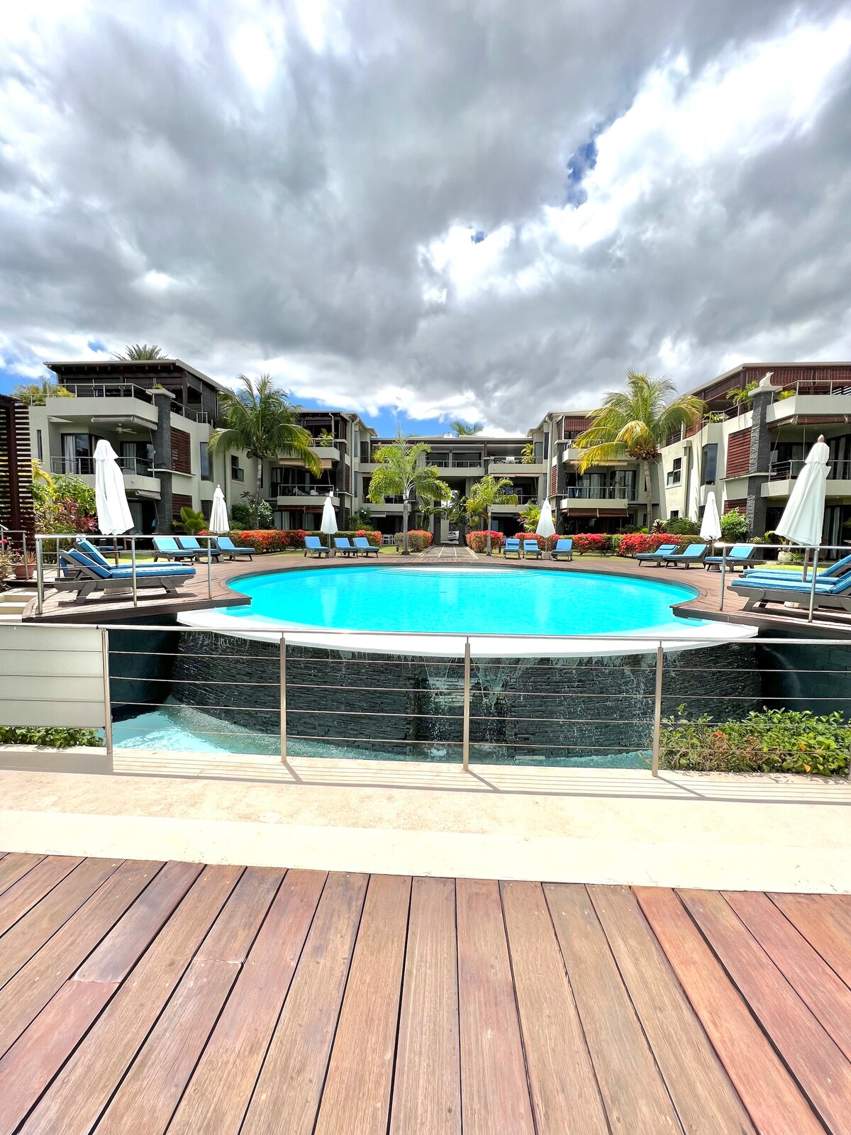A circular pool is surrounded by lounge chairs and umbrellas, set within a landscaped area. Tropical plants enhance the setting, while modern apartment buildings frame the scene in the background under a partly cloudy sky.