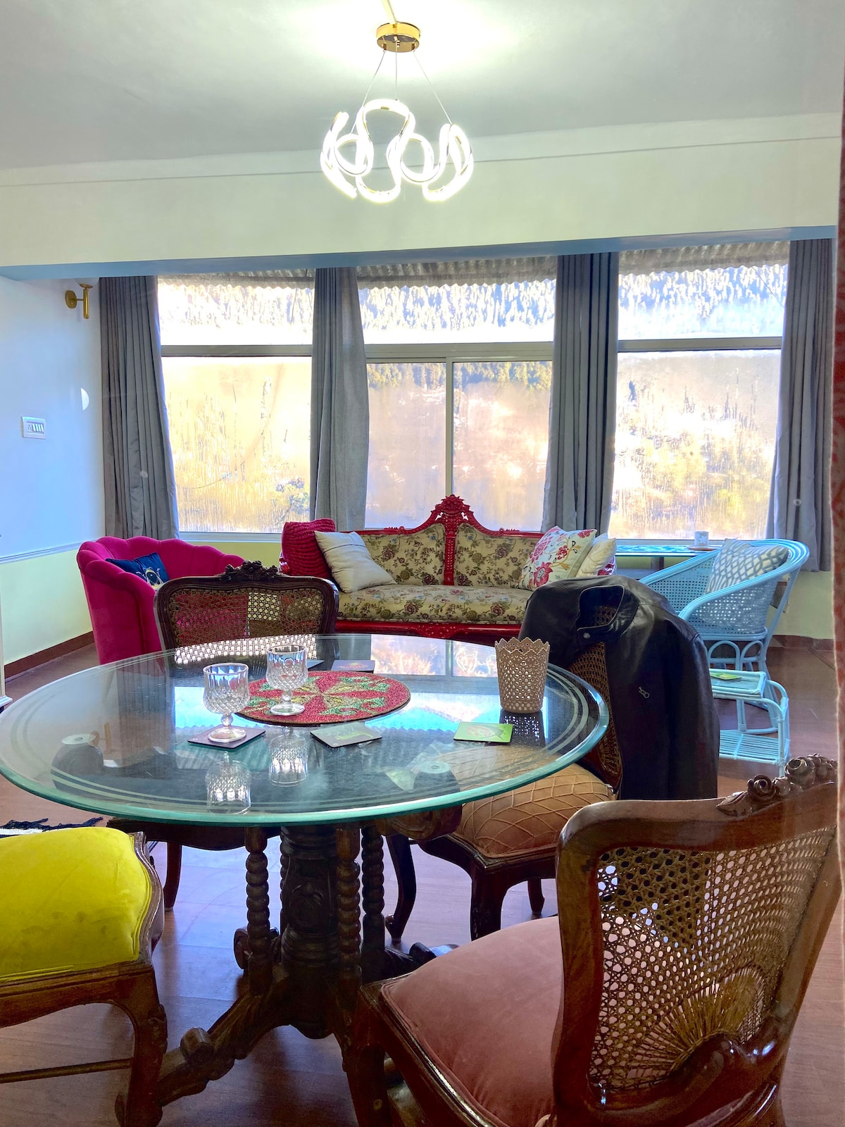 A cozy living area features a round glass dining table surrounded by assorted vintage chairs, including a pink chair and a blue rattan chair. Natural light filters through large windows draped with sheer curtains, highlighting colorful cushions and floral decor on the red sofa.