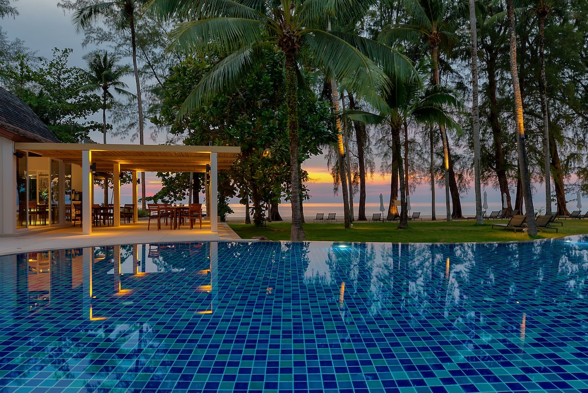 An outdoor swimming pool reflects the vibrant colors of the sunset, surrounded by lush palm trees. Comfortable seating areas and dining tables are visible under a covered pavilion, enhancing the serene beachfront scenery.
