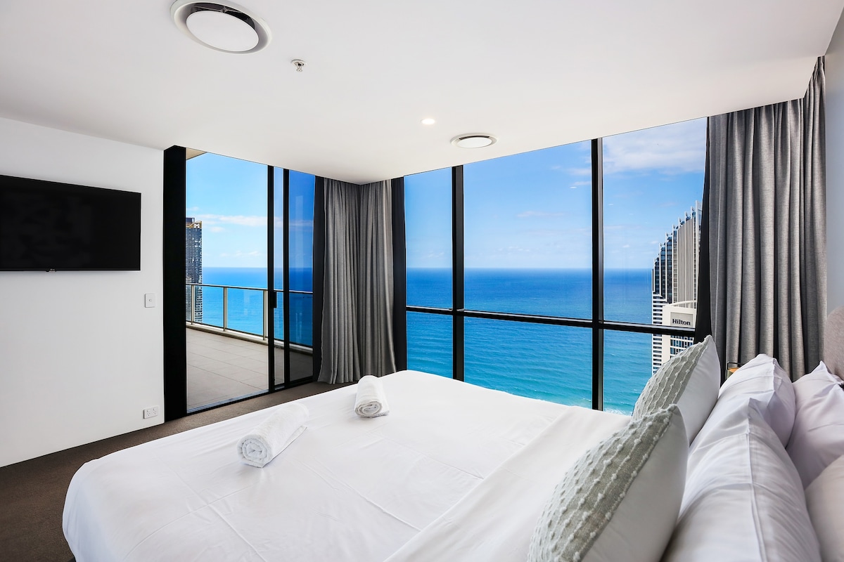 A spacious bedroom features a large window offering panoramic ocean views. A comfortable bed is centered in the room, with neatly arranged towels placed on the bed. Natural light brightens the area, creating a serene atmosphere.