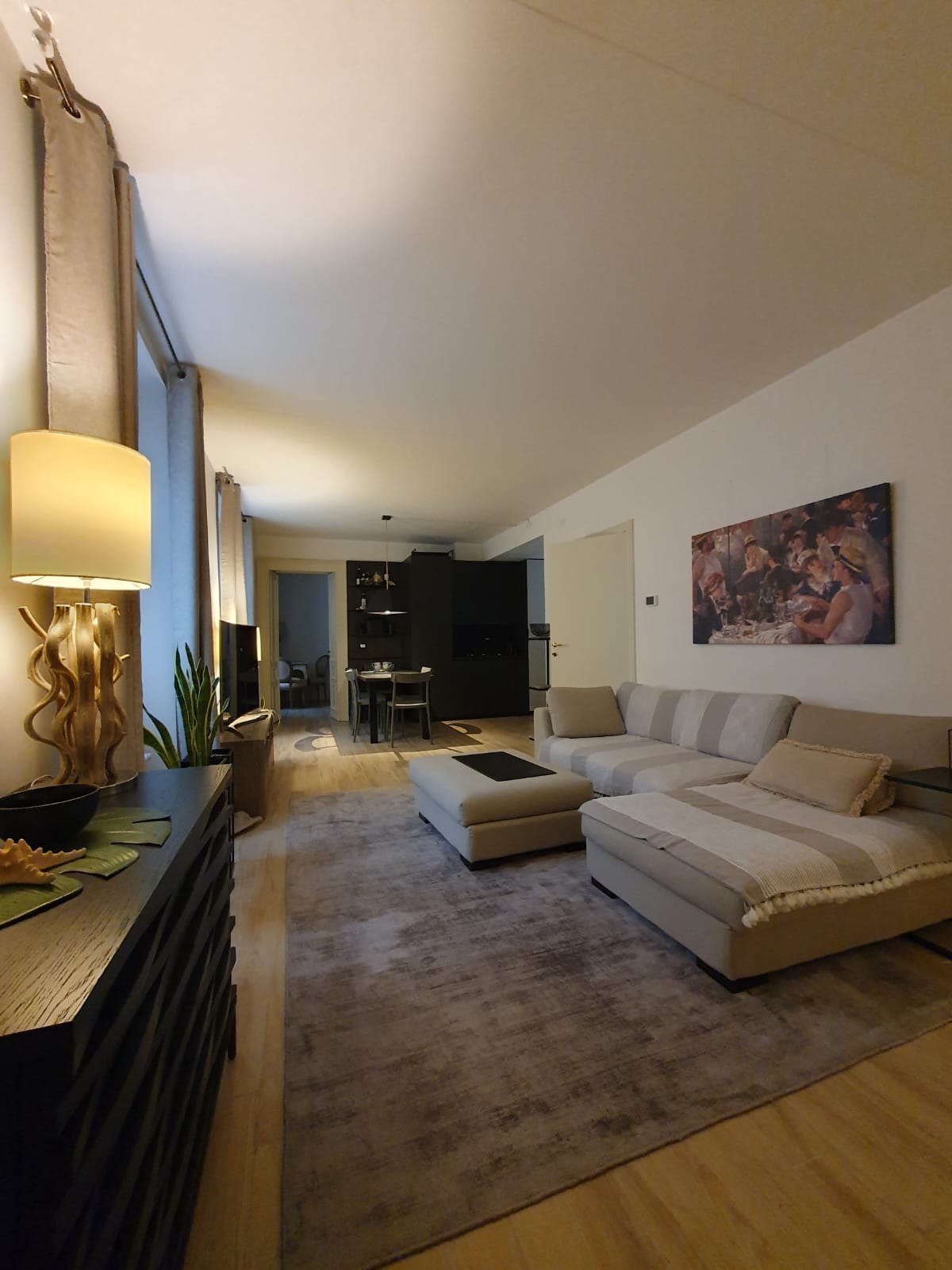 An open-plan living area features a light-toned sectional sofa and a decorative lamp. A rug covers the wooden floor, while a piece of wall art enhances the space. A dining area with chairs is visible in the background, alongside kitchen elements.