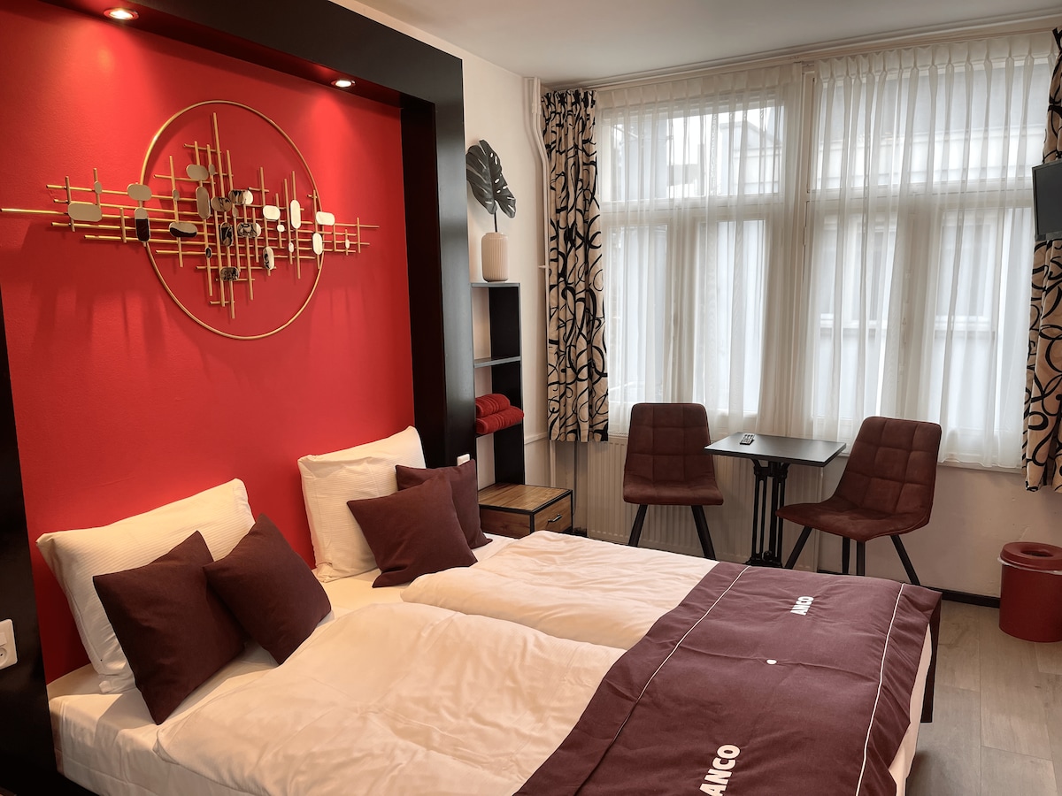 A bright sleeping area features two spacious beds with white linens and burgundy accents. A decorative wall piece in silver and red enhances the backdrop, while natural light filters through sheer curtains. A small dining table and two chairs are arranged near the window.