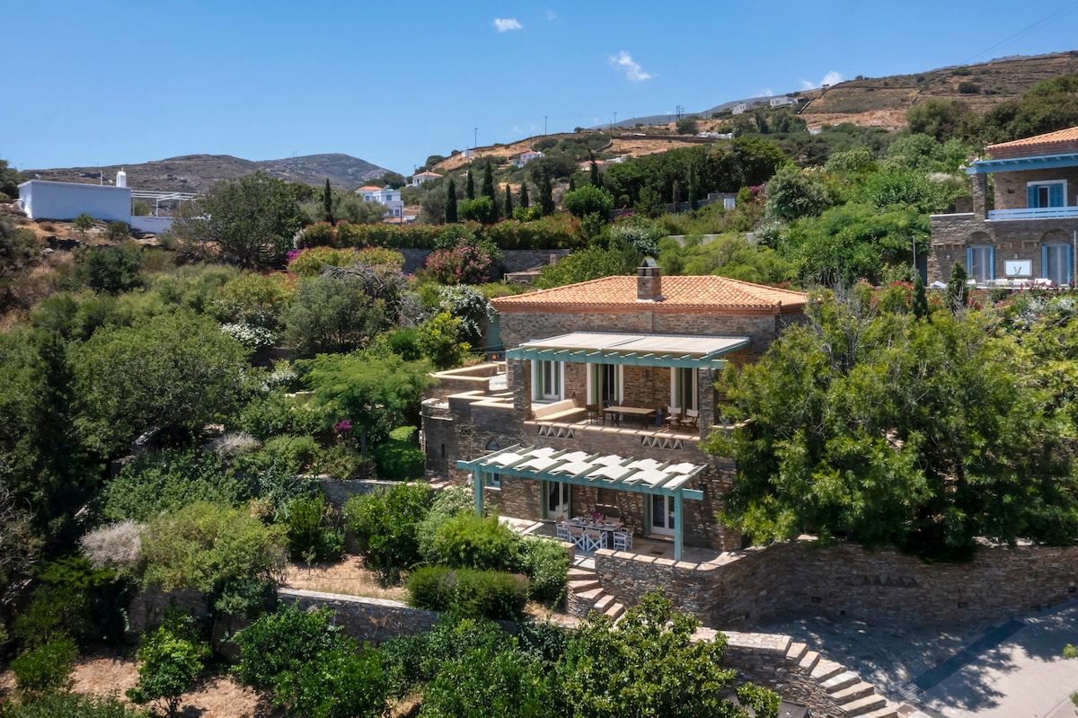 A charming stone villa is set amidst lush greenery and colorful flowering plants. The structure features a terracotta roof and large windows, allowing natural light to fill the interior. Outdoor seating is present on the porch, providing a serene spot to enjoy the landscape.