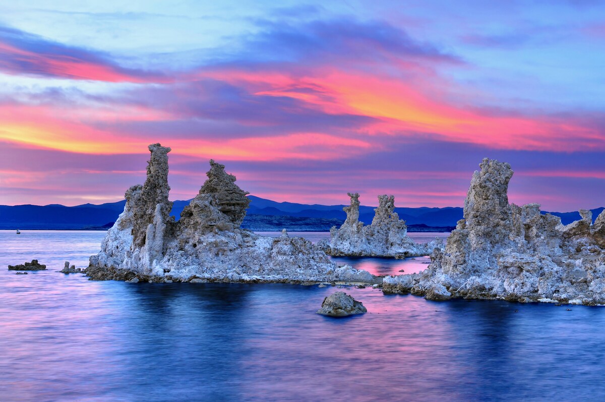 A serene landscape features unique rock formations emerging from tranquil water, with colorful shades of pink and purple in the sky at sunset. The distant mountains provide a gentle backdrop, while the smooth water reflects the evening hues.